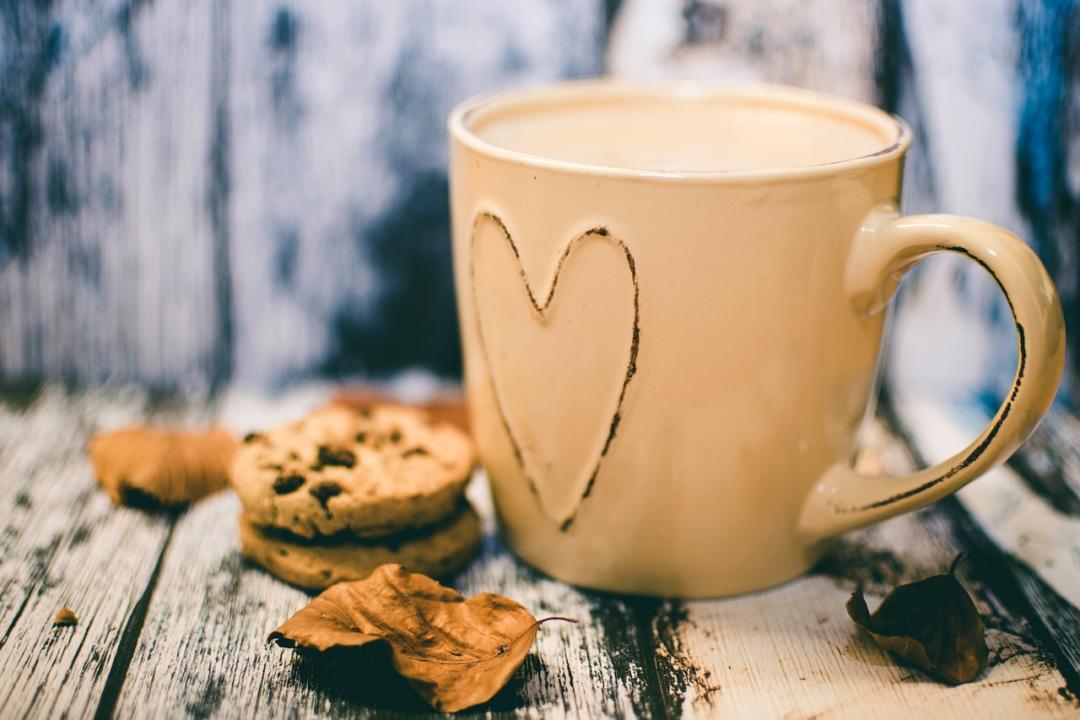 Photo of a mug with a love heart on it filled with coffee next to some leaves and cookies on a wooden backdrop